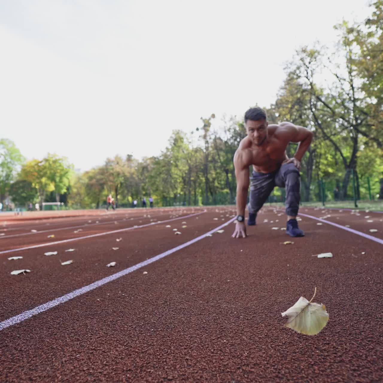 Sportive runner on outdoor athletics track. Athletic man without shirt running on stadium track on green trees background. Motion camera around.
