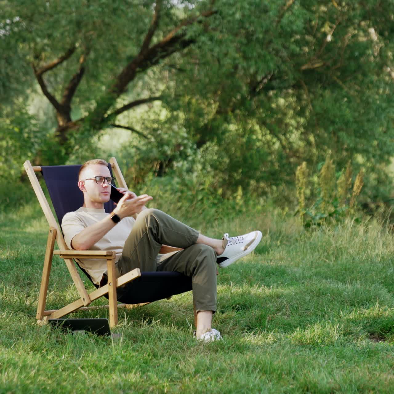 Relaxed male speaks on the phone sitting in the folding chair. Remote work conversation in the woods