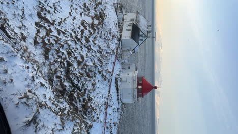 Vertical-handheld-shot-from-inside-of-a-helicopter-as-it-circles-a-small-lighthouse-on-a-rocky-snow-covered-island-at-sunset