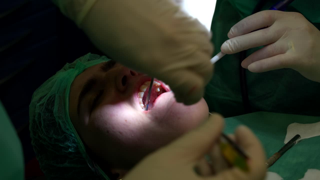 dentista poniendo puntos de sutura en la boca sangrienta de su paciente después de la cirugía de injerto y el asistente lo limpia con una gasa