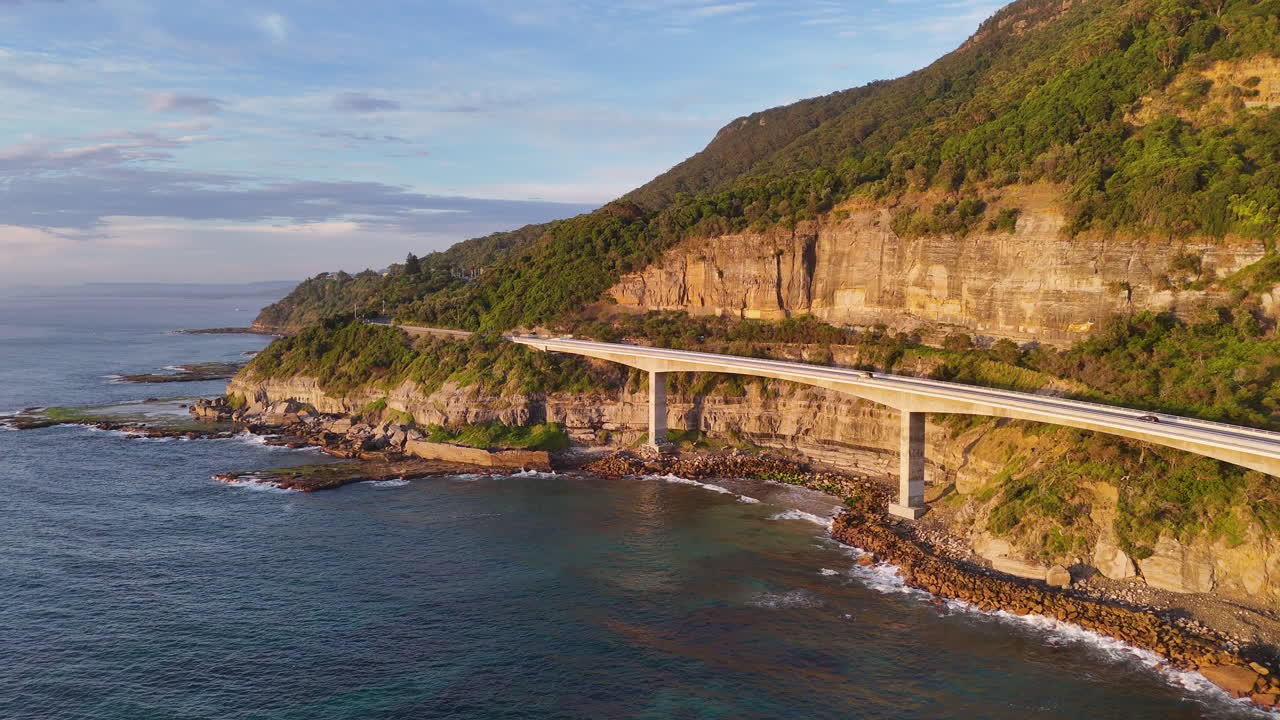 Aerial footage of the Sea Cliff Bridge, with a vehicle navigating the curving road above the blue waters of the Pacific. Drone footage.