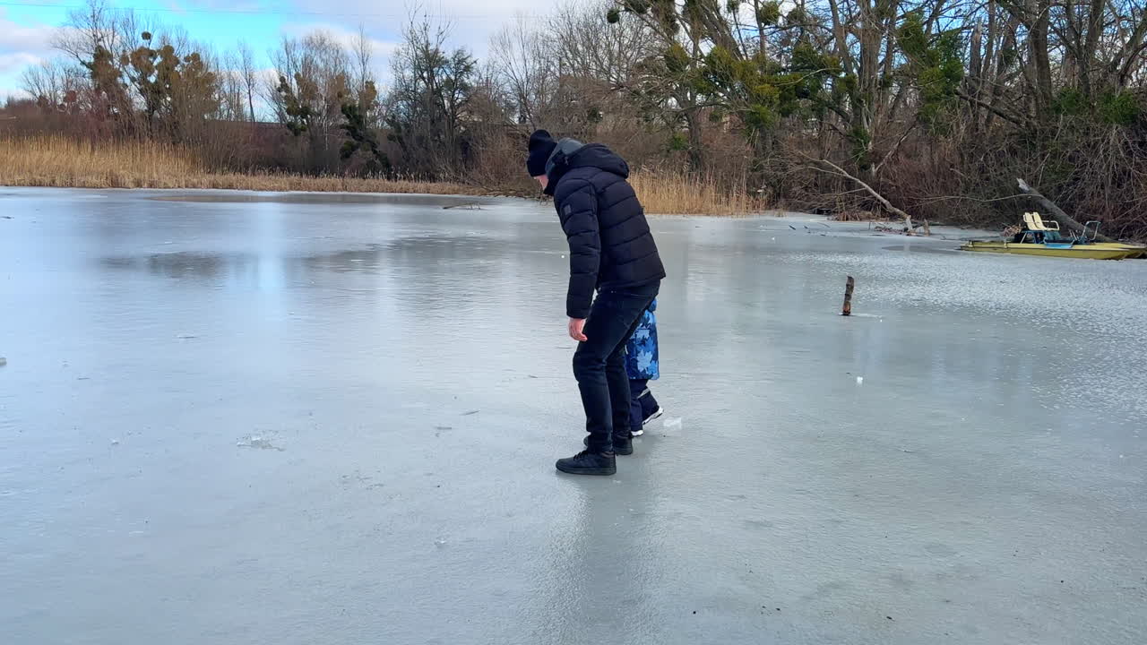 Man and a toddler boy are walking by the frozen river. Dad and son sliding by the ice in winter.