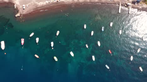 aerial top down view flying sideways across fishing boats in the mediterranean sea with crytal clear blue water and beach in santorini, greece