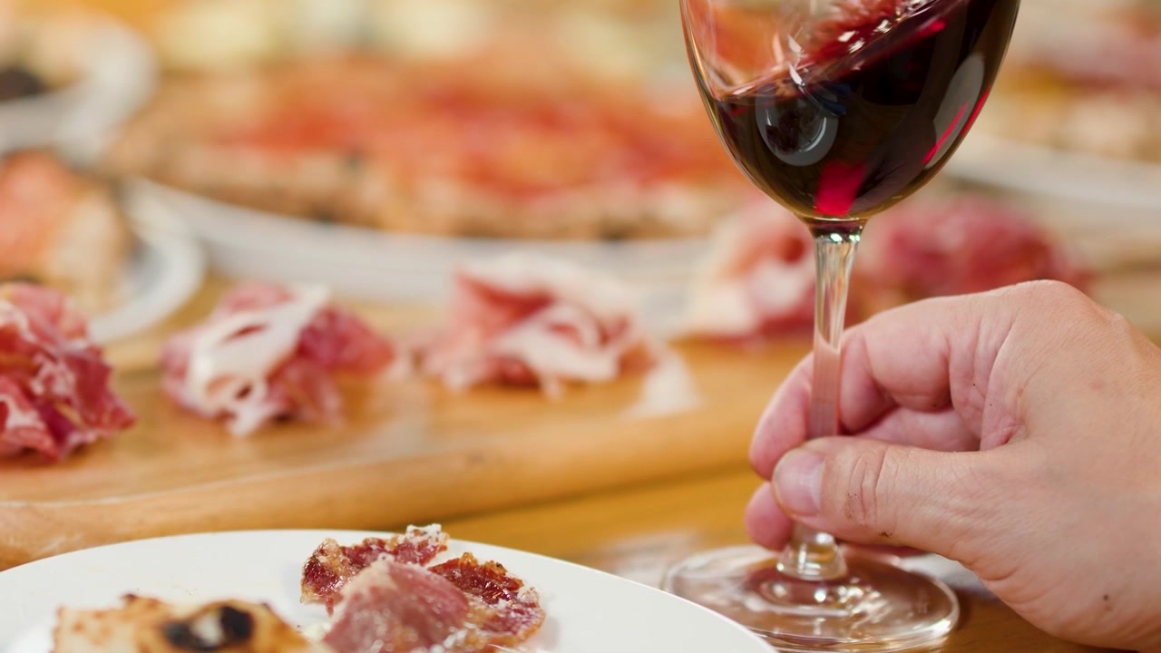 Una mano regge un bicchiere di vino rosso su un tavolo con pizza e salumi