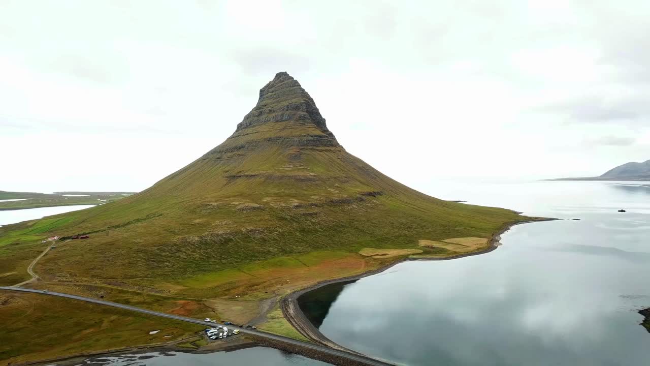 Kirkjufell, a striking mountain in Iceland, rises beside calm coastal waters and lush green fields, framed by dramatic natural landscapes, offering an iconic and picturesque view