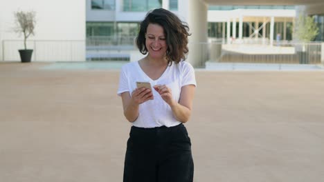 Hermosa-Mujer-Sonriente-Caminando-Con-Un-Teléfono-Inteligente-En-La-Calle.