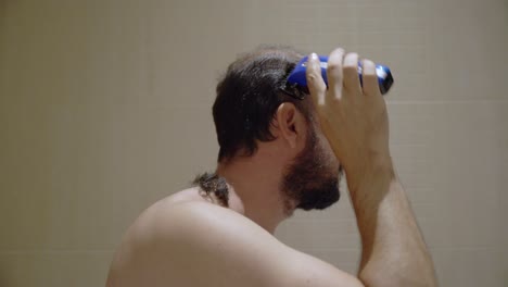 A shirtless man cuts his own hair at home in the bathtub in bathroom with an electric clipper shave during lockdown quarantine isolation