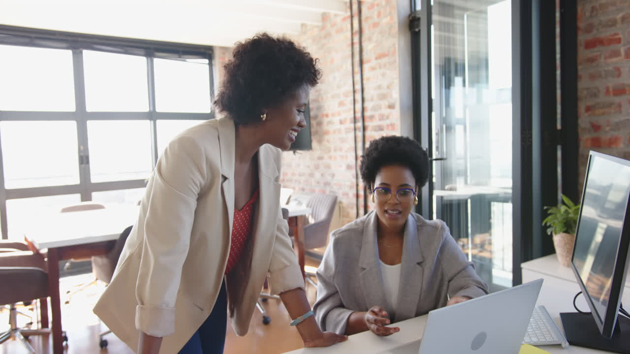 Discussing project, two businesswomen collaborating at desk with computer in office