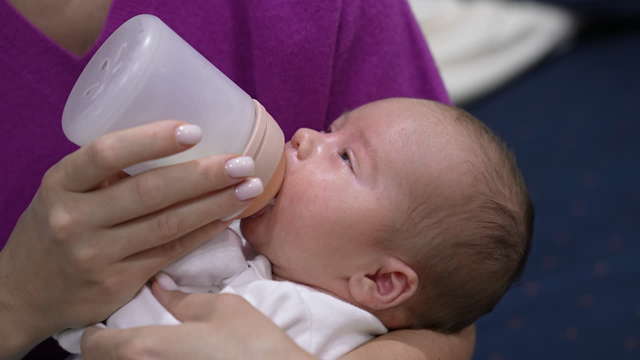 Mother is holding a child in her arms and feeding a baby from a bottle. Baby eating milk and falling asleep gradually. Close up.