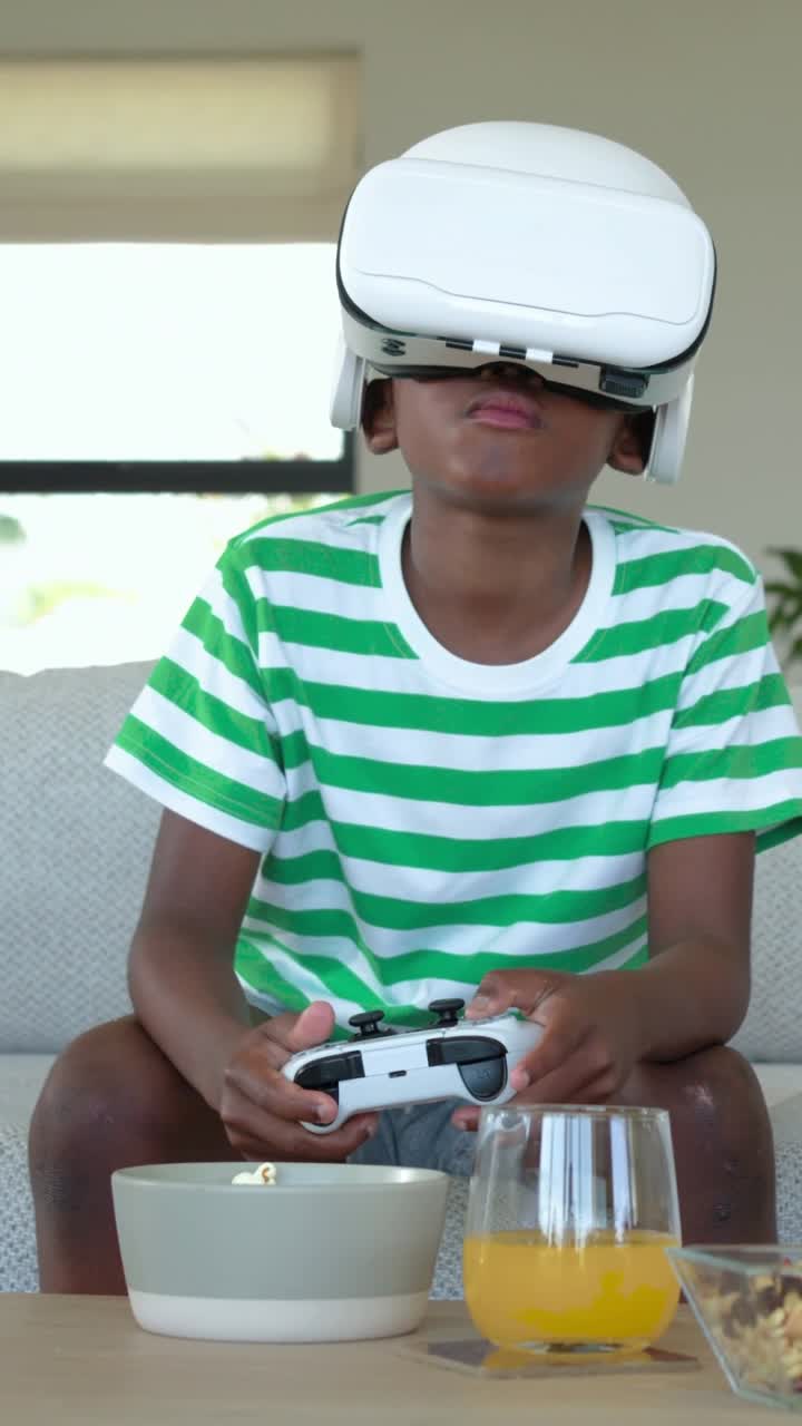 Video verticale: bambino che gioca in maglietta con visore VR, usando un controller sul divano del soggiorno mentre il gioco inizia.