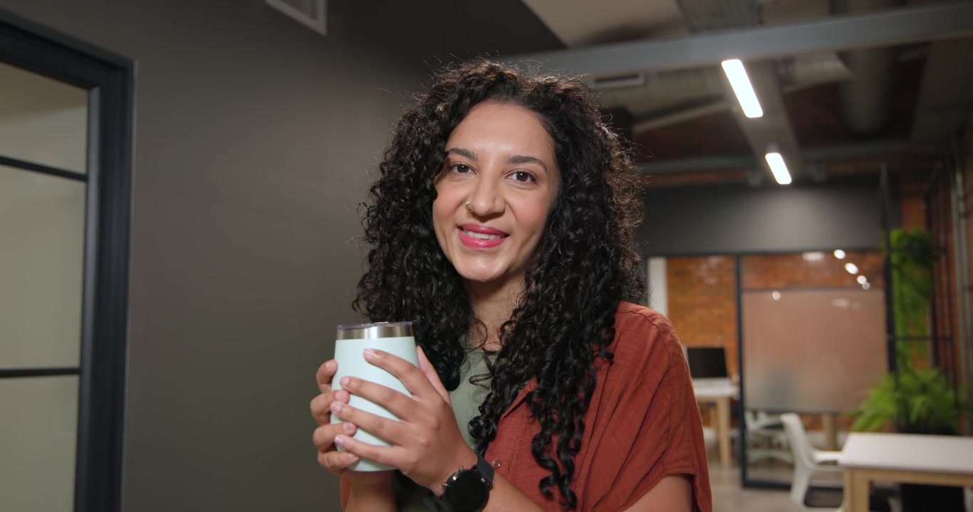 Mulher afro-americana com cardigã cor ferrugem, de pé no escritório, segurando caneca térmica, fazendo uma pausa e sorrindo