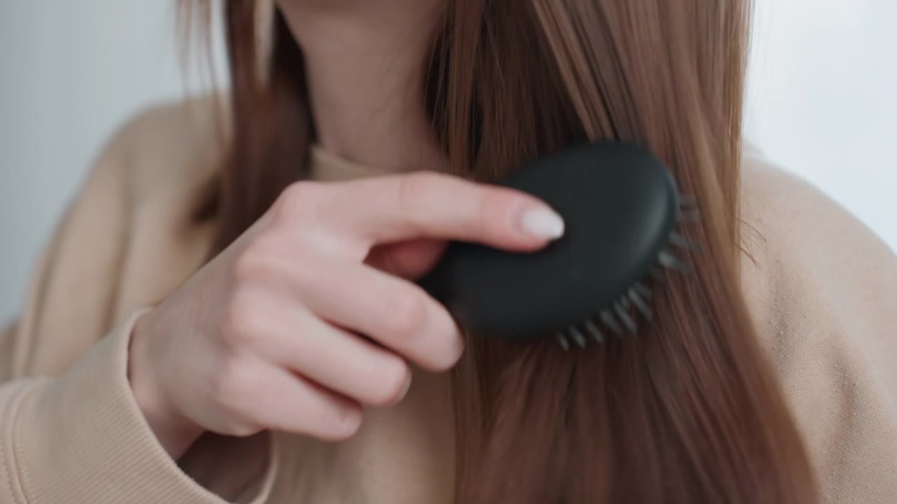 Caucasian woman preparing hair. Closeup of woman smoothing her hair. Focused woman gently brushing brown hair strands. Calm woman in neutral clothing grooming her hair for coaching session