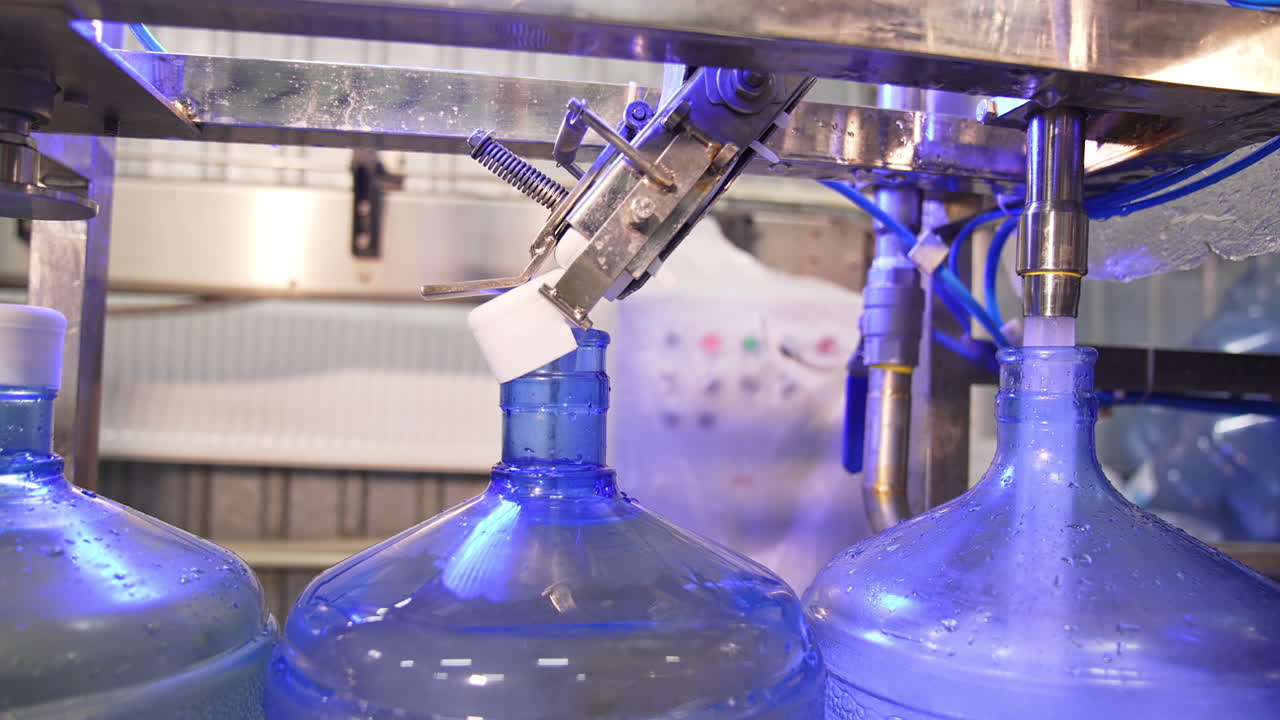 Conveyor belt with big blue flasks moving slowly. Bottles are filled with clean mineral water and being corked.