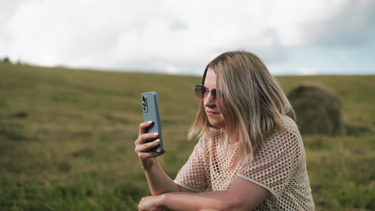 mujer en cuclillas en vastas tierras de cultivo operando un teléfono con un insecto posado en su mano, entorno rural al aire libre con paisajes naturales, balas de heno en la distancia