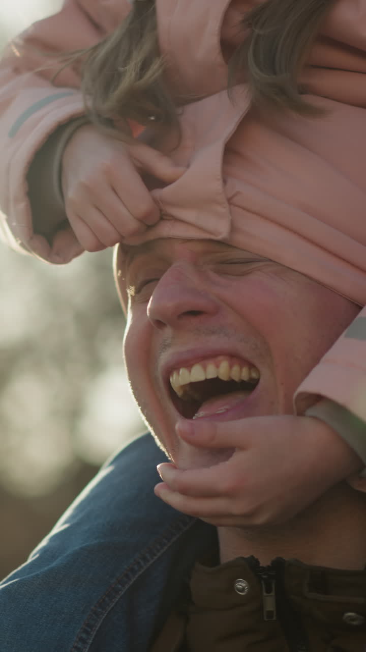 A little girl in a pink cap and jacket sits on her dad's neck, with his head snugly tucked in her jacket. The girl playfully pulls the jacket off his head while they both smile brightly