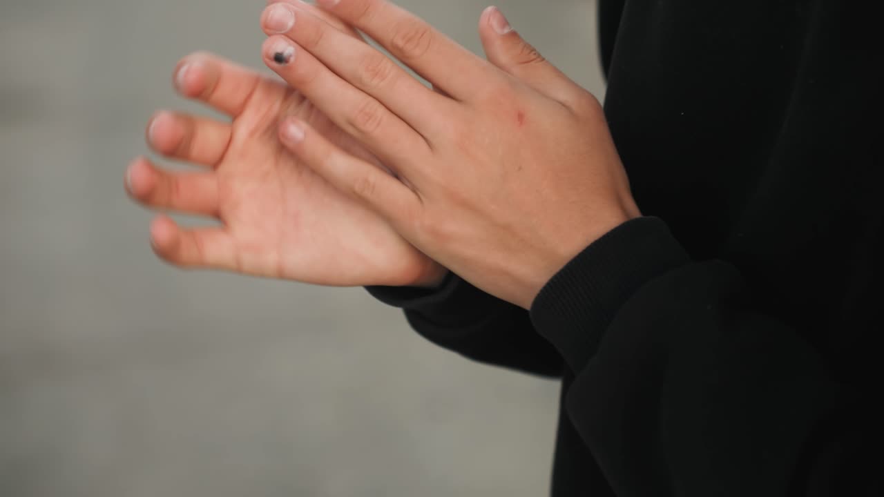 Si esibisce con vitalità sul marciapiede cittadino. Una persona con la felpa esegue gesti vivaci di spettacolo di strada. Mani che si strofinano con energia contro la felpa mentre l'artista si prepara in un contesto urbano di cemento.