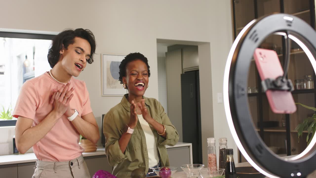 Diverse co-hosts greeting viewers via ring light smartphone at kitchen island, synchronizing waves