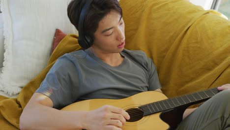 asian boy wearing headphones playing guitar lying on bean bag at home