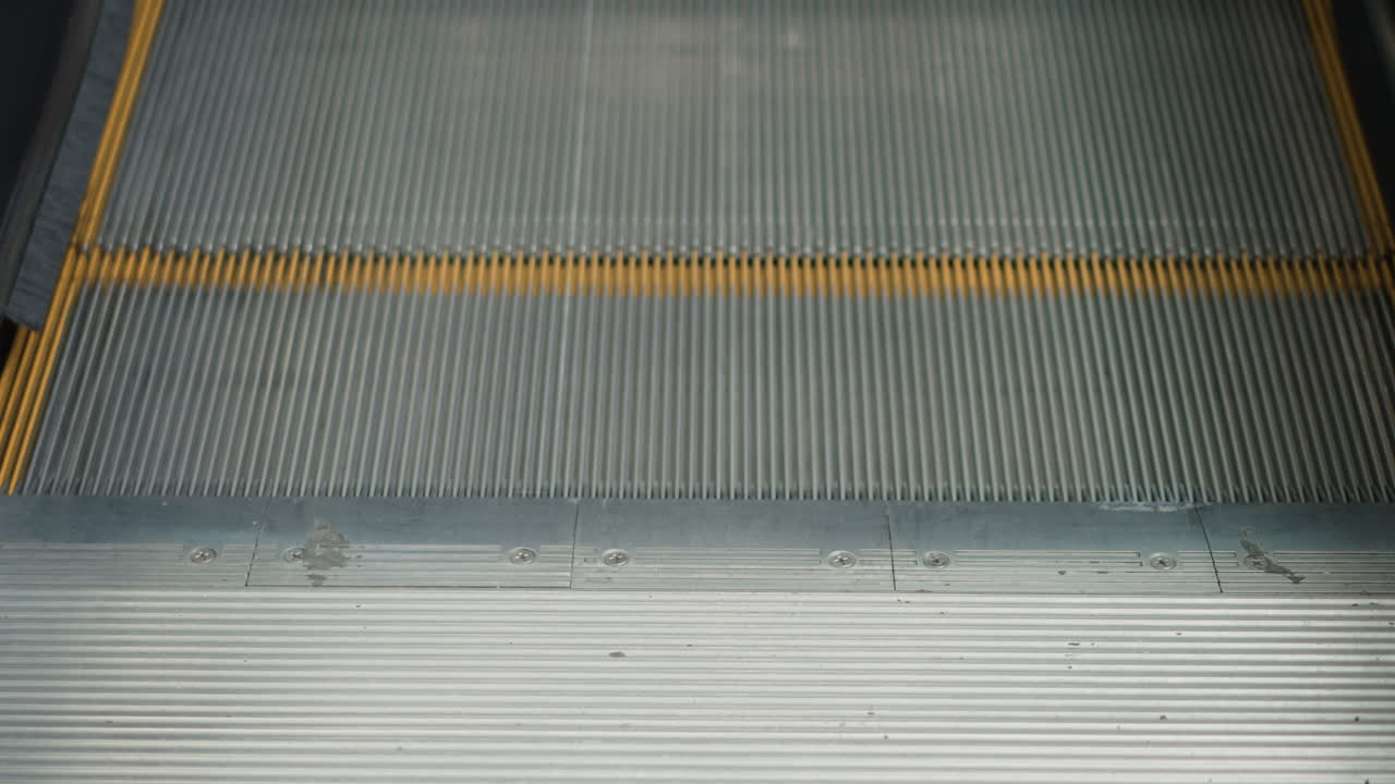 close up view of moving walkway metal steps with yellow safety edges and wear marks, focusing on textured ridged surface and contrasting transition area in modern shopping mall environment