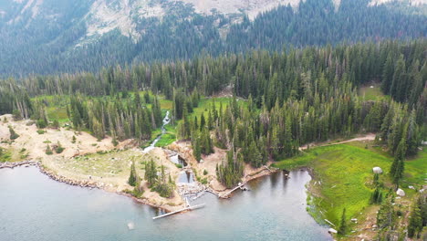 aerial drone circling beaver damn in clear blue lake surrounded by thick pine tree forest in nederland colorado rocky mountains with flowing creek water