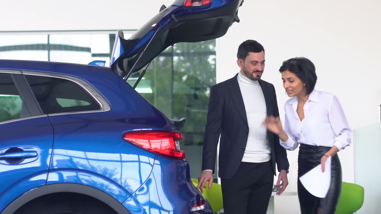 una vendedora de automóviles profesional le está hablando a un comprador interesado sobre un coche de lujo con el maletero abierto en una exposición de automóviles.