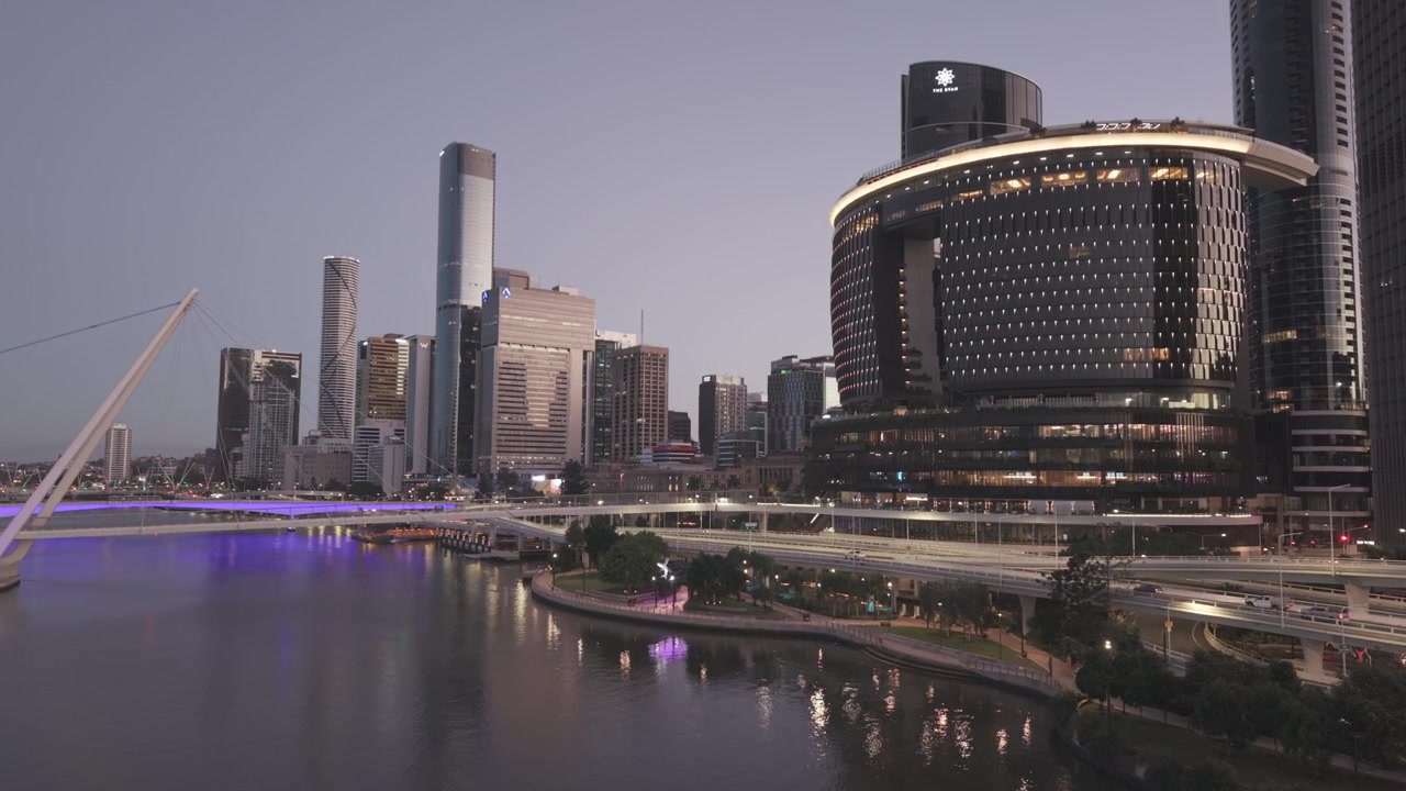 A close aerial view shows the Brisbane skyline and the Neville Bonner Bridge glowing above the river at dusk.
