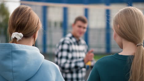two ladies in cyan and green sweaters standing on volleyball court, watching coach in plaid shirt instruct them while holding volleyball under arm, background slightly blurred