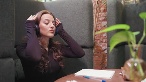 light skin lady with blond hair wearing black headphones, seated in cozy booth with eyes closed, holding book on table beside notepad and pen, surrounded by warm decoration and soft lighting ambiance
