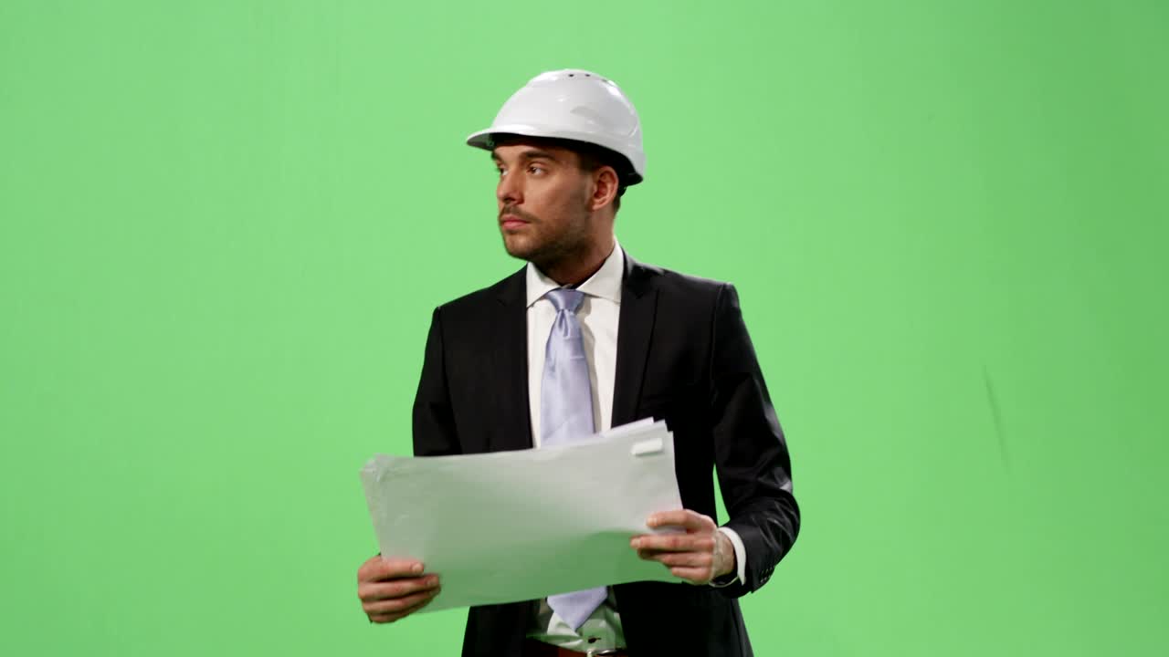 un hombre de negocios con un casco y un traje está caminando y mirando documentos de papel en una pantalla verde de maqueta en el fondo.