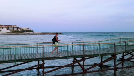 Seitenansicht-Eines-Jungen-Mannes-Mit-Rucksack-Und-Sonnenbrille,-Der-Auf-Einem-Holzsteg-Am-Robinson-Beach-An-Der-Bulgarischen-Schwarzmeerküste-Läuft