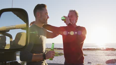 Happy-caucasian-gay-male-couple-standing-by-car-drinking-bottles-of-beer-on-sunny-day-at-the-beach