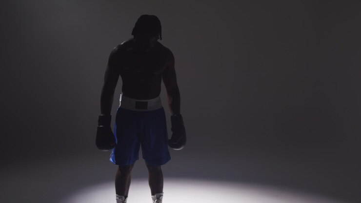 Silhouetted Studio Shot With Low Key Lighting Of Boxer Wearing Gloves Standing In Pool Of Light Against Grey Background