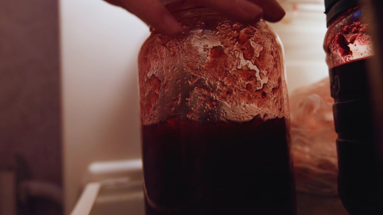 Jar of red jam in fridge, home cook stores homemade preserve in glass jar on shelf dim interior light, condensation on lid, plasticwrapped leftovers beside jar, rustic kitchen mood and closeup texture