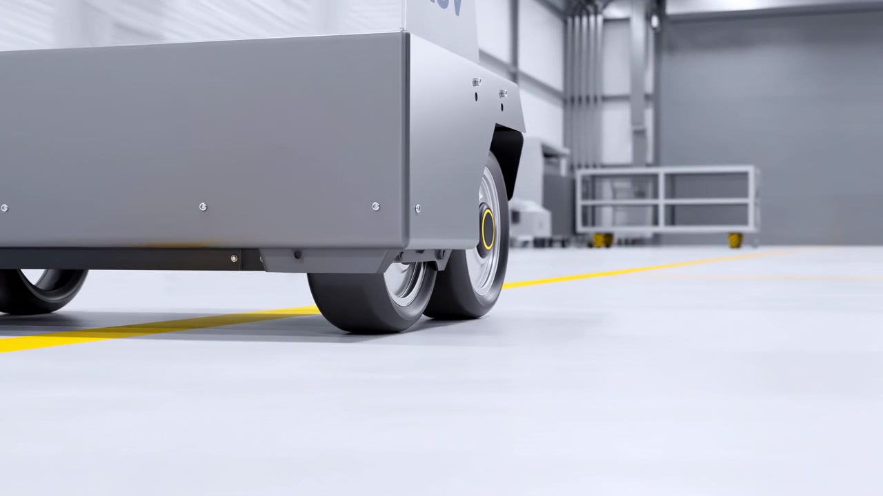 Small robotic vehicle rolls forward across bright indoor floor as wheels rotate beneath compact body, showing smooth motion, clean surfaces, and industrial surroundings with soft lighting.