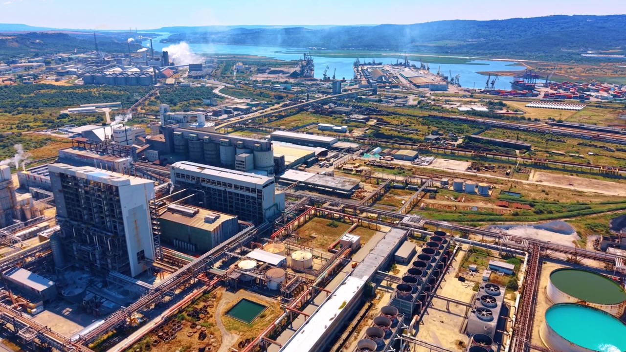 Aerial view of industrial complex with cooling towers. High angle shot shows factory buildings, round water pools, port, smoke, green fields, hills.