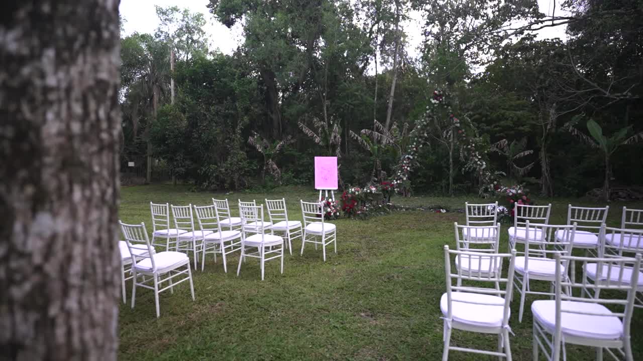 Right-tracking scene reveals outdoor wedding ceremony aisle with chairs and decoration, surrounded by lush trees and vegetation