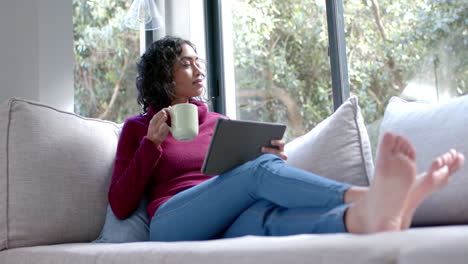 Young-Asian-woman-relaxes-on-a-couch-at-home,-holding-a-tablet-and-a-mug