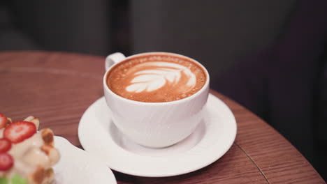 close-up view of latte with leaf-shaped milk foam art in white cup on saucer next to waffles topped with cream and strawberry slices, all set on brown wooden table in cozy environment