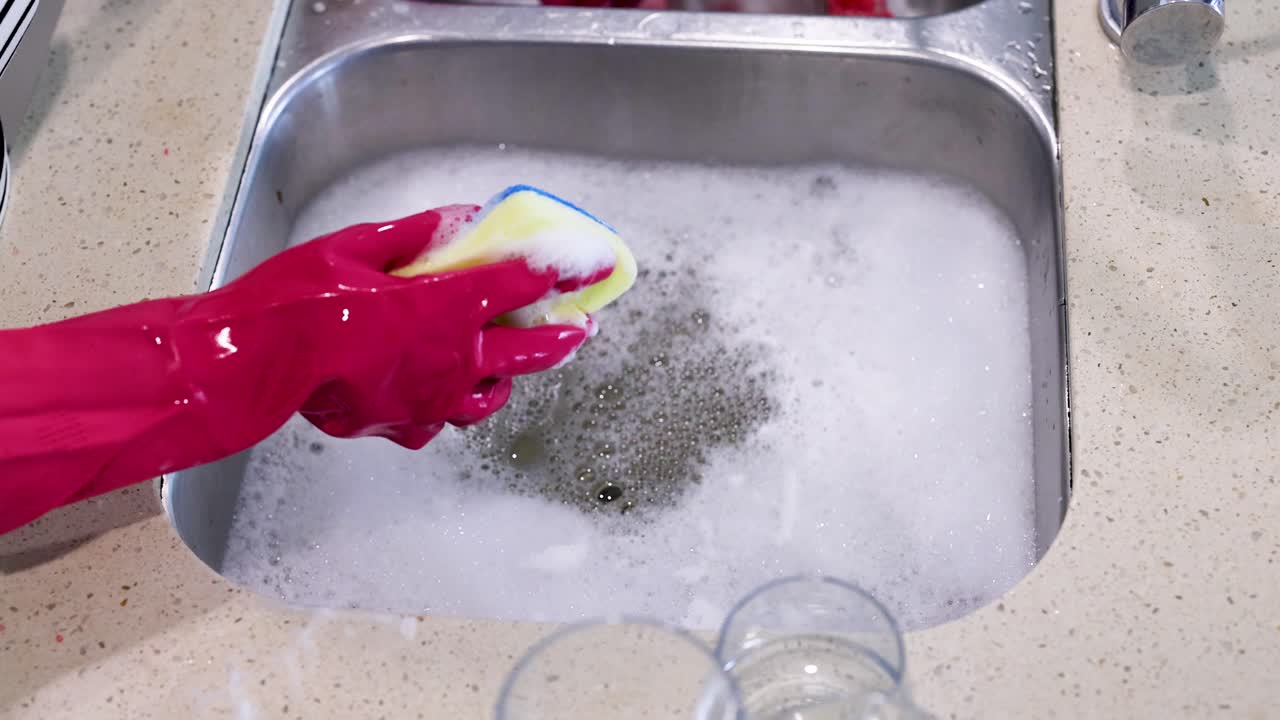 Hands in pink gloves clean a colander in a kitchen sink filled with soapy water, under bright lighting