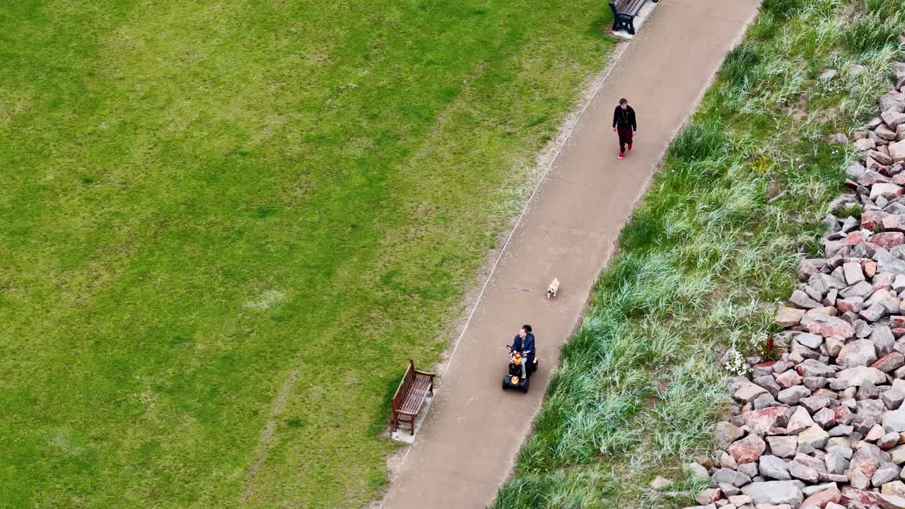 Elderly person on mobility scooter, dog walker, and benches along Carnoustie coastal walkway, daylight aerial