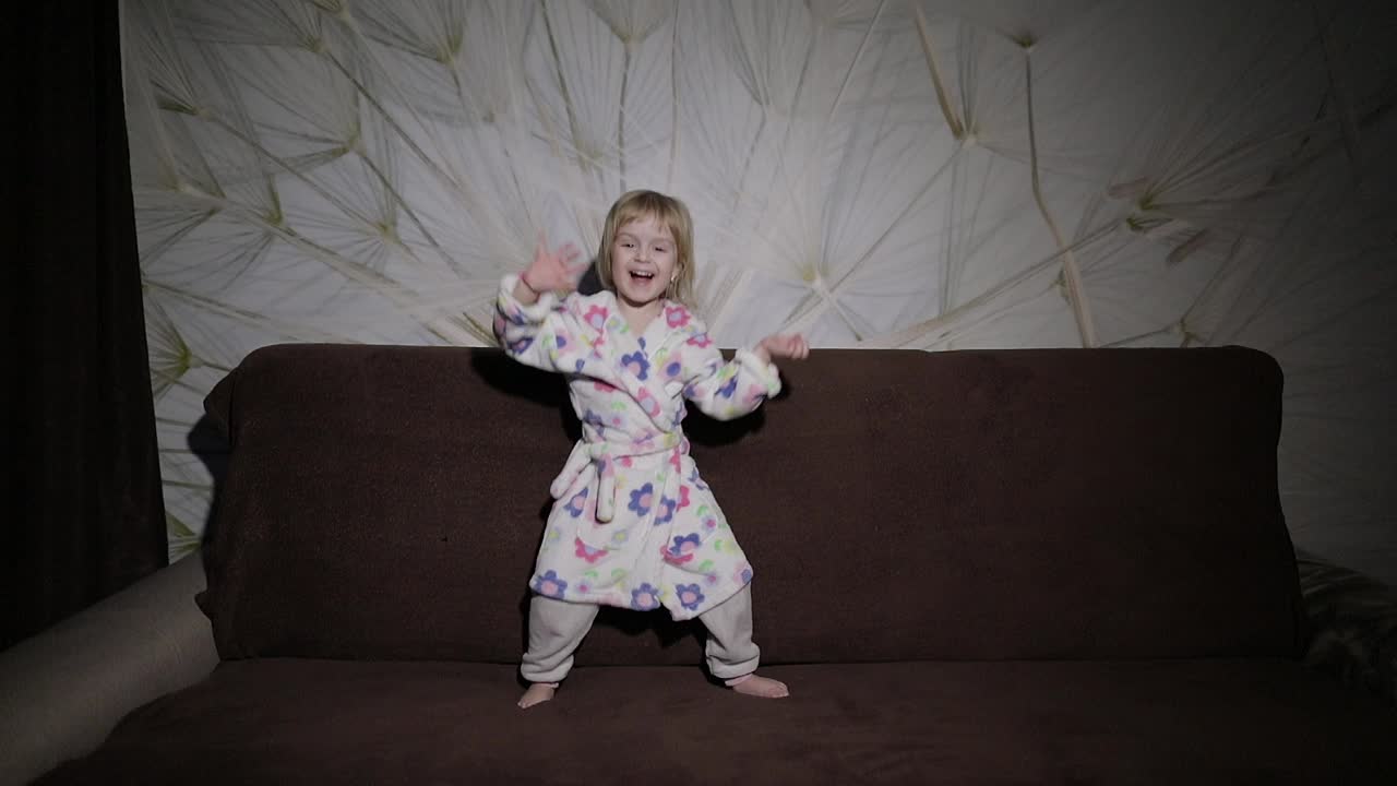 Little cute girl with blond hair jumps on sofa. Bathrobe clothes. Happy