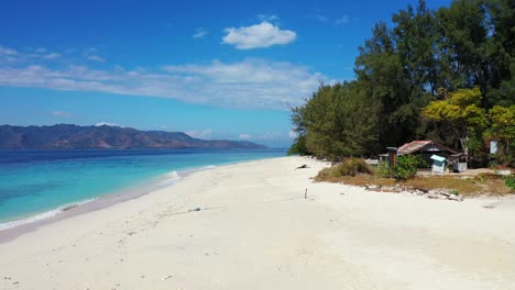 Pristine-exotic-beach-with-white-sand-under-shadow-of-tall-trees,-washed-by-blue-azure-sea-on-a-bright-sky-over-mountains-background-in-Bali