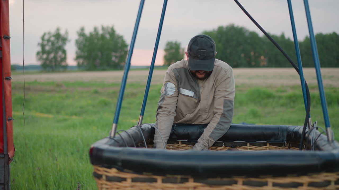man hands gas cylinder to woman inside wicker balloon basket as duo prepare burner assembly and inspect pressure gauge on roadside under cloudy sky with red envelope backdrop before flight launch