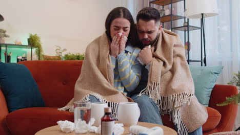 sick ill family man woman suffering from cold sitting on home couch sneezes wipes snot into napkin
