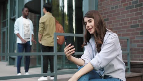 caucasian student woman having a video call on phone in the street near the college