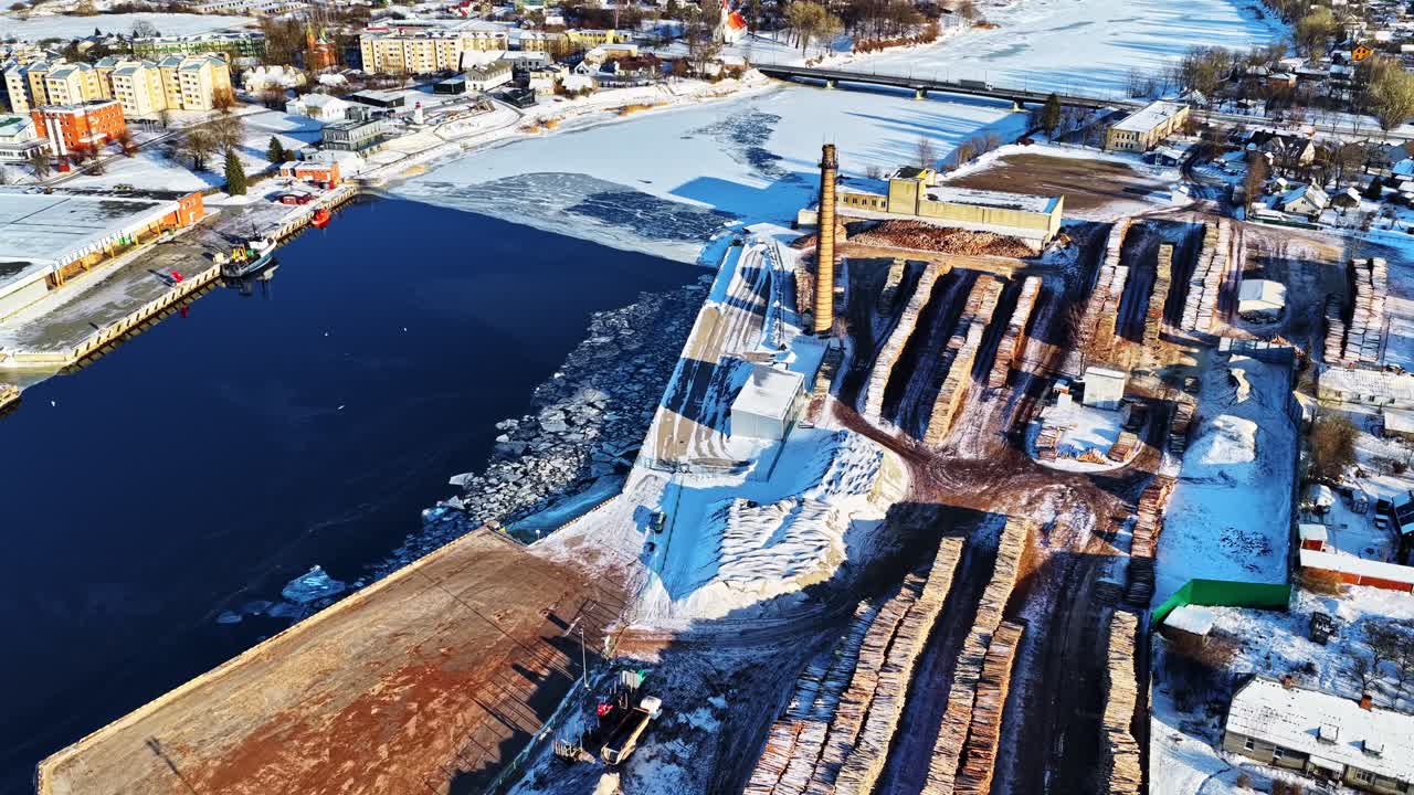 Salacgrīvan satama esittelee talvitoimintojaan: jäätyneen joen varrelle pinottuja tukkeja ja paikallista kaupunkia ilmasta käsin aurinkoisena talvipäivänä.