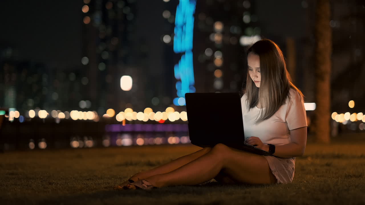 una mujer desarrolladora de software se sienta en un parque en una noche de verano en la ciudad y escribe código mirando la pantalla de una computadora portátil. imprime el artículo en una computadora portable. blogger responde a las publicaciones
