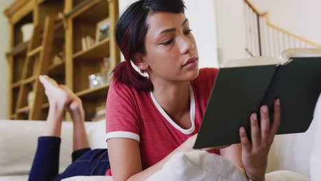 Happy-biracial-teenager-girl-sitting-on-sofa-and-reading-book,-in-slow-motion