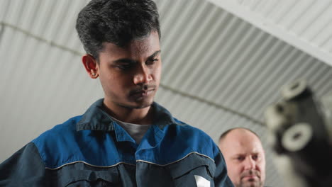 automotive engineers in workshop, one in blue uniform focused on task, while colleague observes closely, mechanical equipment and tools fill background, creating an industrial atmosphere