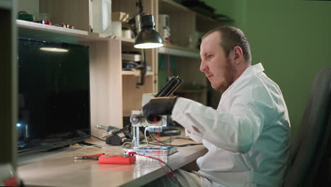 una vista de cerca de un técnico con una bata blanca de laboratorio y guantes negros prueba cuidadosamente un circuito bajo un microscopio usando un voltímetro en un laboratorio bien organizado